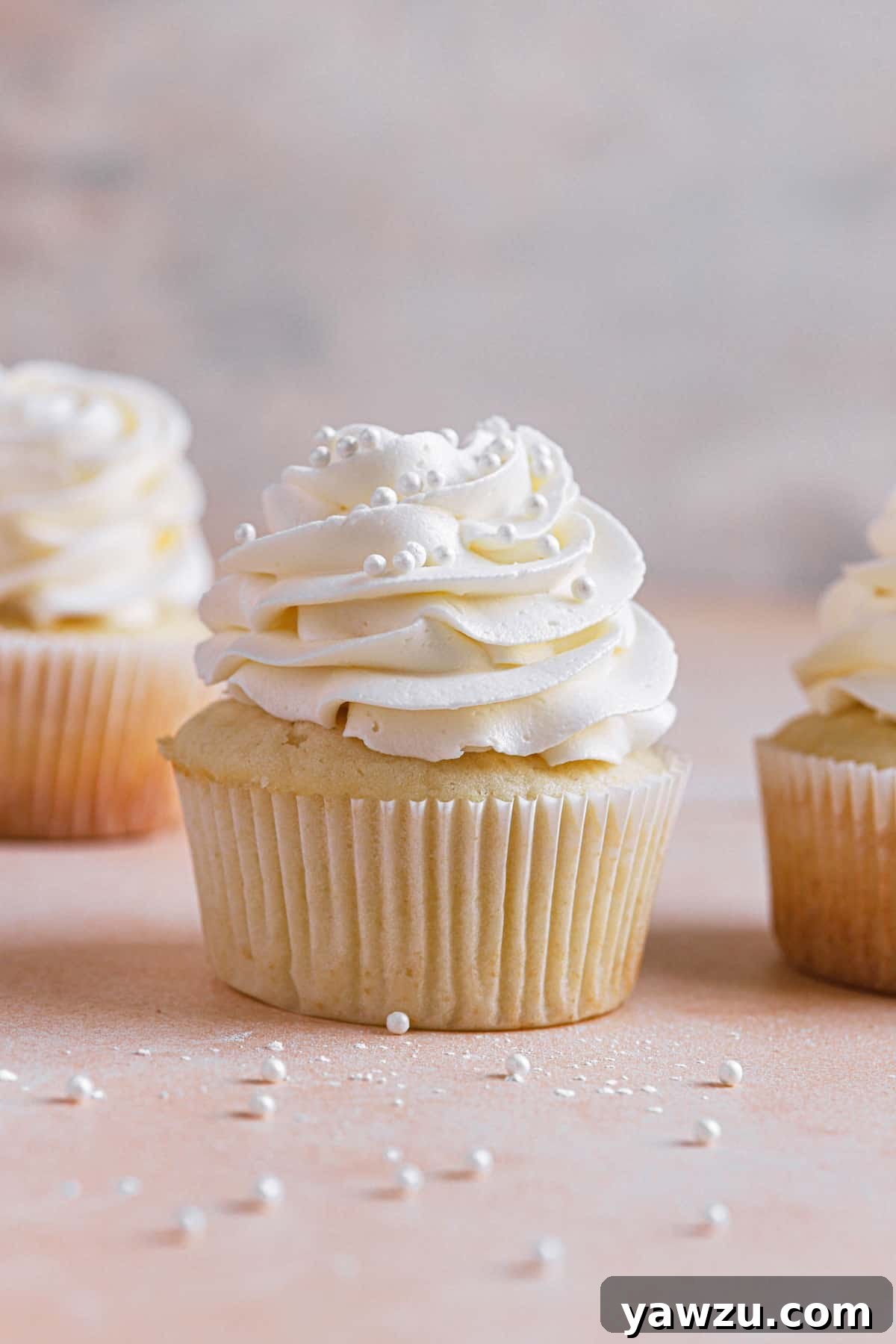 A vanilla cupcake with vanilla frosting and sprinkles in the middle another to the right and one in the back left.