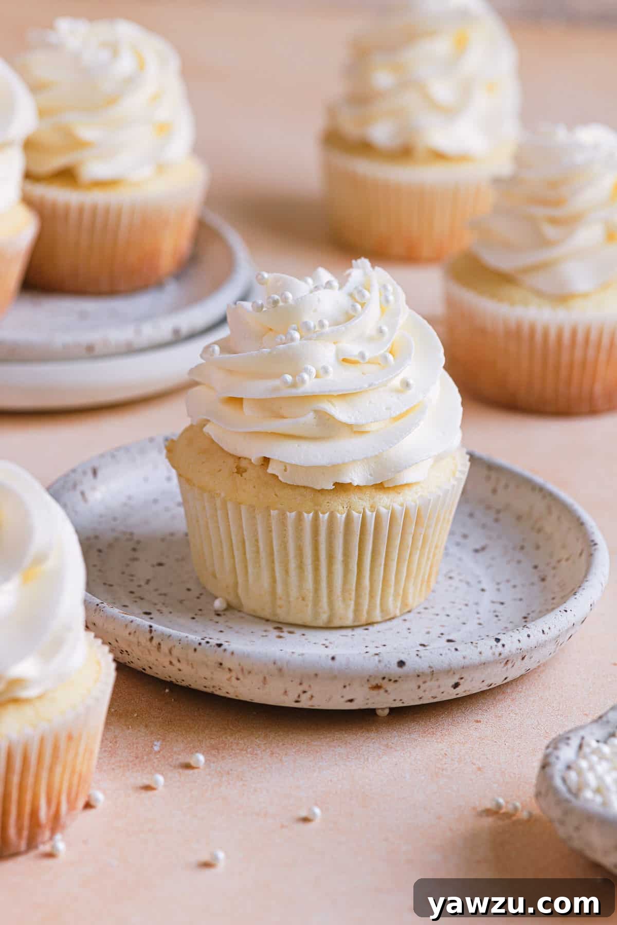 A white speckled plate with a vanilla cupcake in the center and other vanilla cupcakes on the counter in the back.