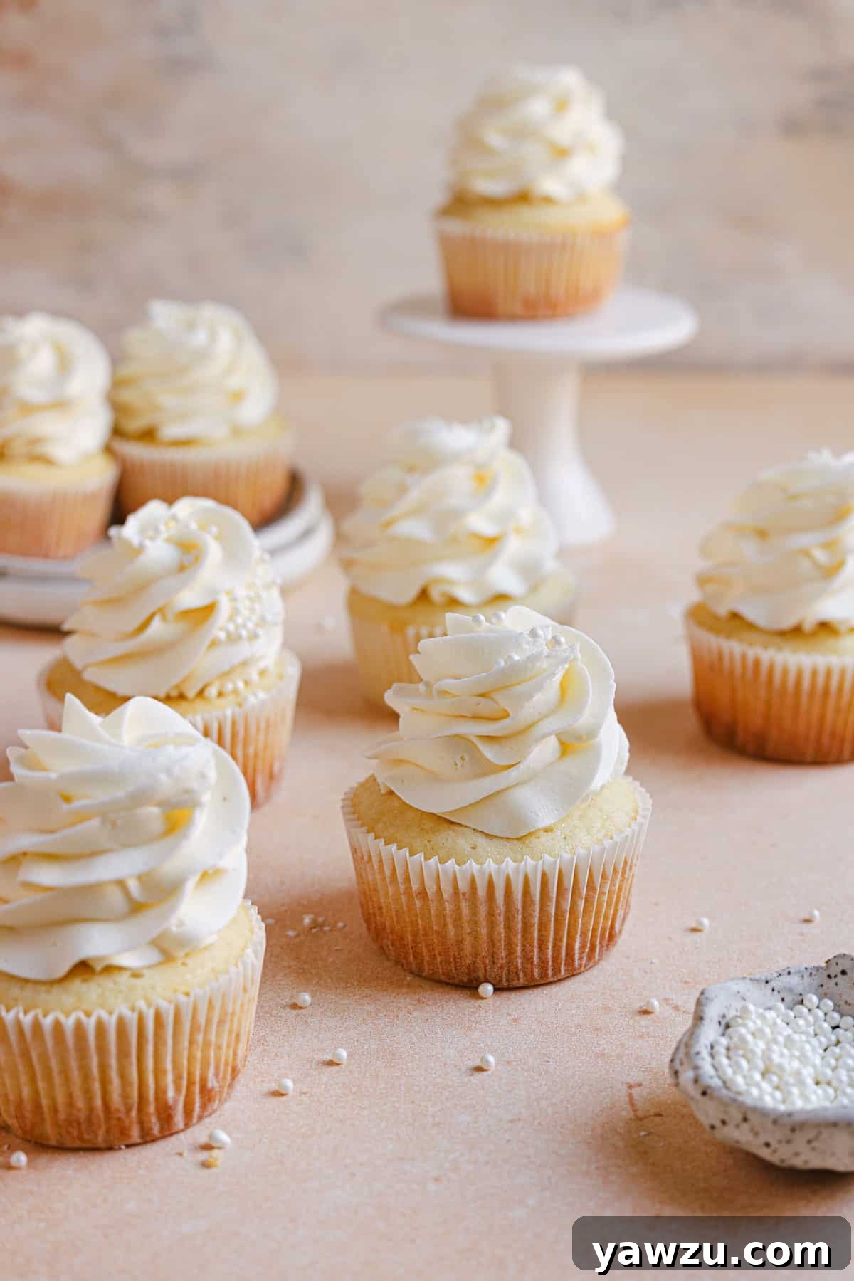 Moist vanilla cupcakes on a counter with a cupcake on a cake stand in the back.