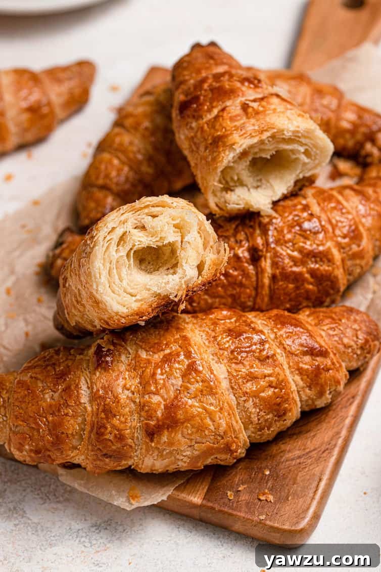 Freshly baked homemade croissants on a wooden cutting board, with one croissant elegantly broken in half to reveal its exquisite flaky layers and airy interior.